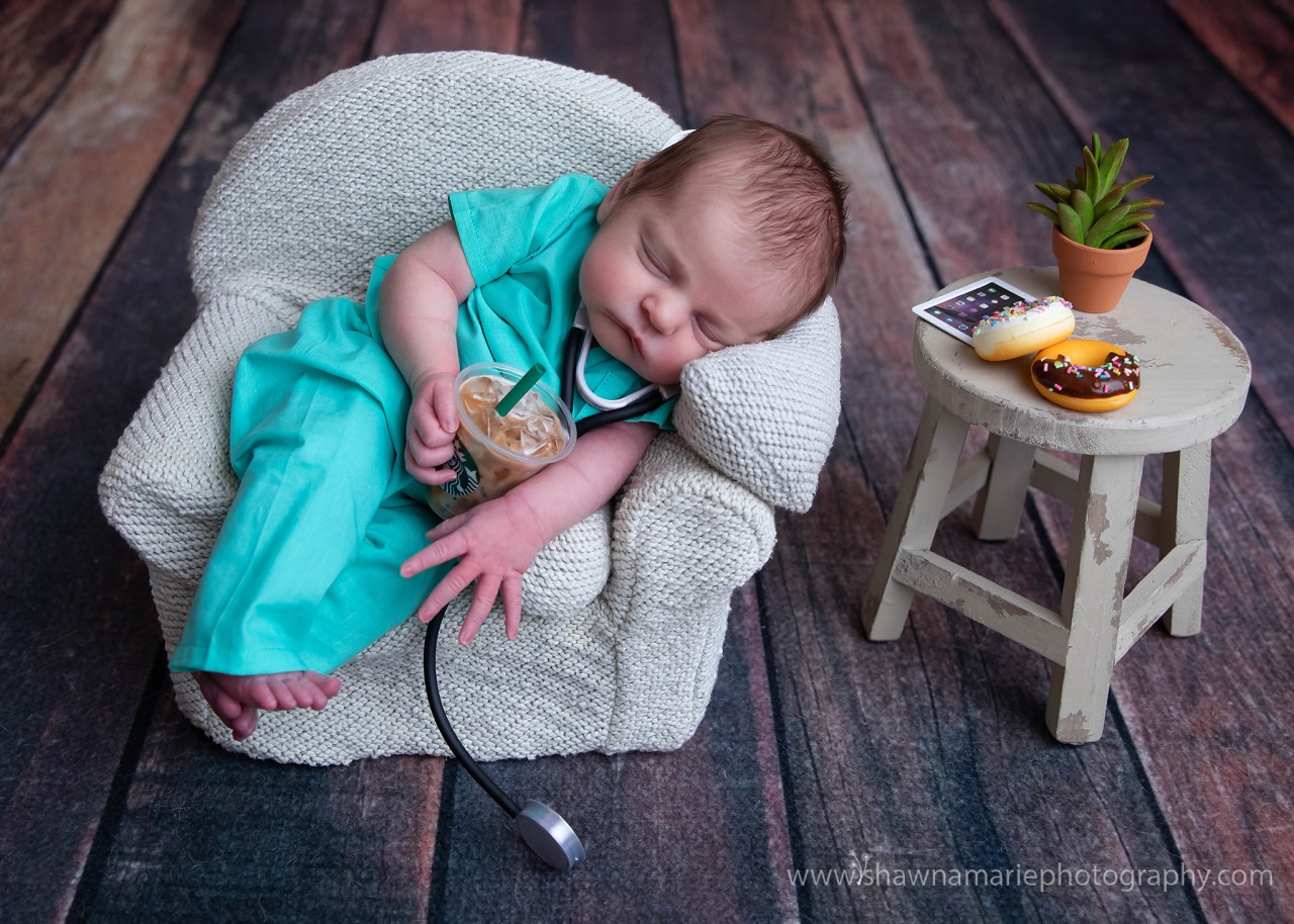 two week old baby Alivia dressed as a nurse holding starbucks and a stethoscope with donuts and an ipad on a table